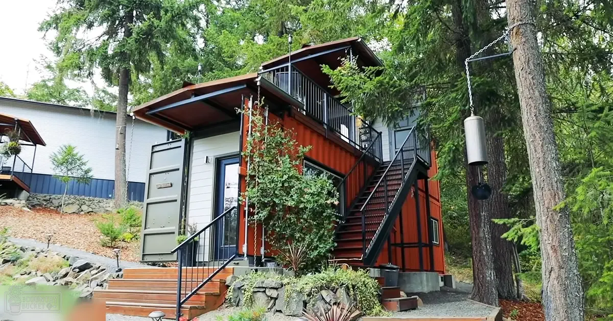 A modern house built from shipping containers with red exterior in a forested area, with a wind chime and staircase leading to the second floor during spring or summer under overcast conditions.