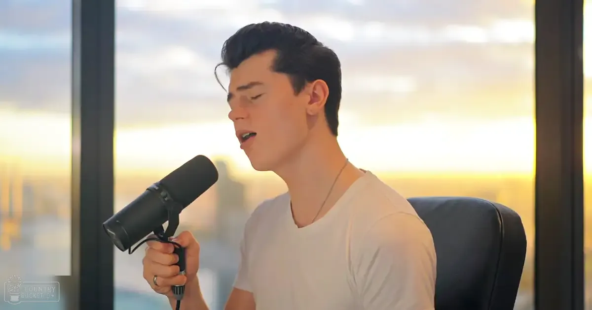A young adult male singer in a relaxed white V-neck T-shirt, holding a professional condenser microphone, with large windows showing a cityscape at sunset.