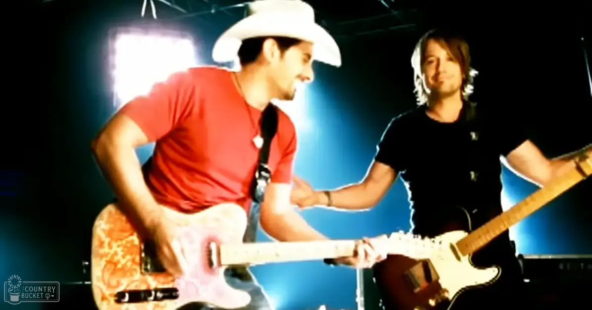 Musical performance scene with two adult male musicians on stage, wearing casual clothing and holding electric guitars under blue stage lights in an indoor venue.