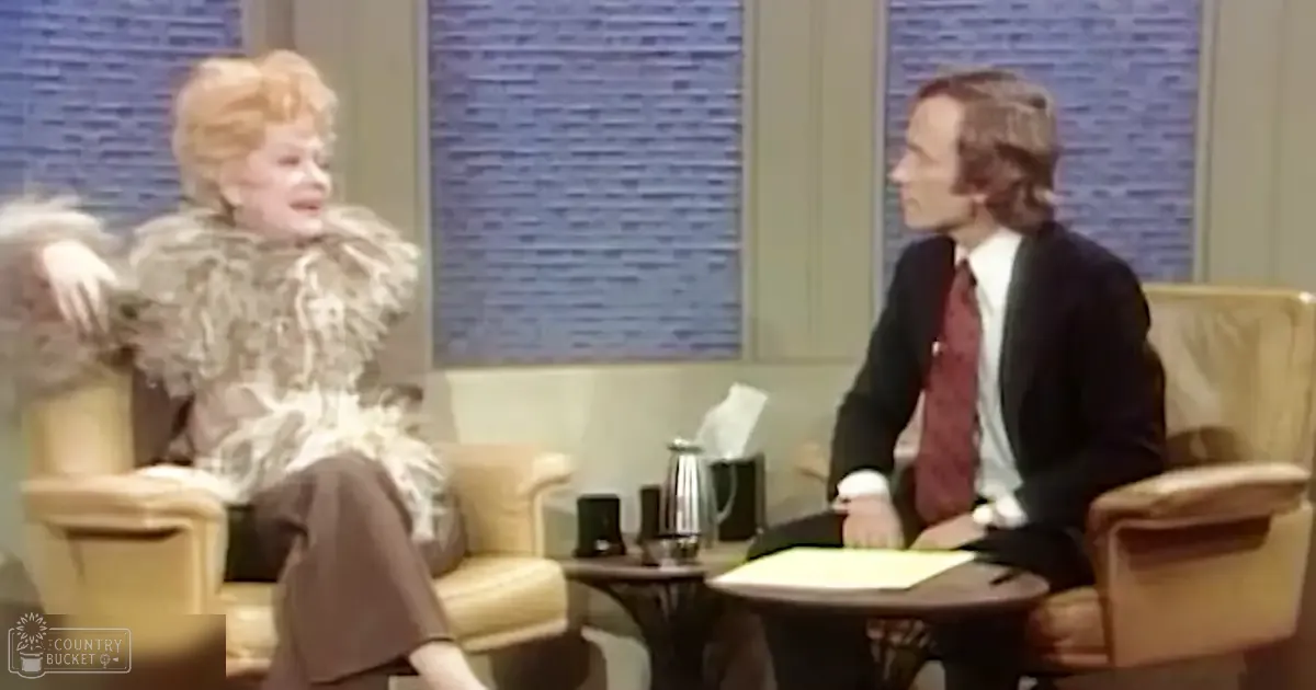 A woman wearing a vintage-style white ruffled top with feather-like details interviews an adult male in a formal dark suit jacket and red patterned tie, set against modern blue textured panels of an indoor studio.