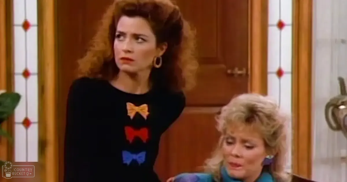 A young adult female in a home setting with wooden furniture, wearing a black top with colorful bow designs and large hoop earrings; another middle-aged woman is present in the background.