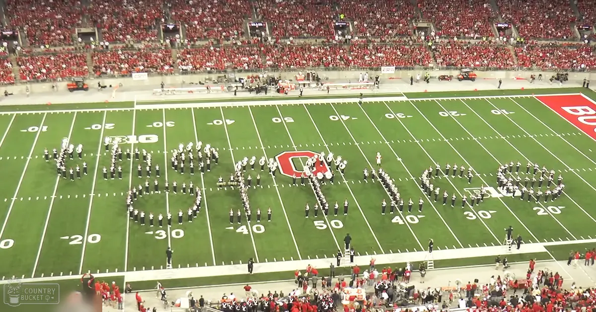 Young adult male marching band performing at a football stadium in fall, red spectator attire and green turf visible.