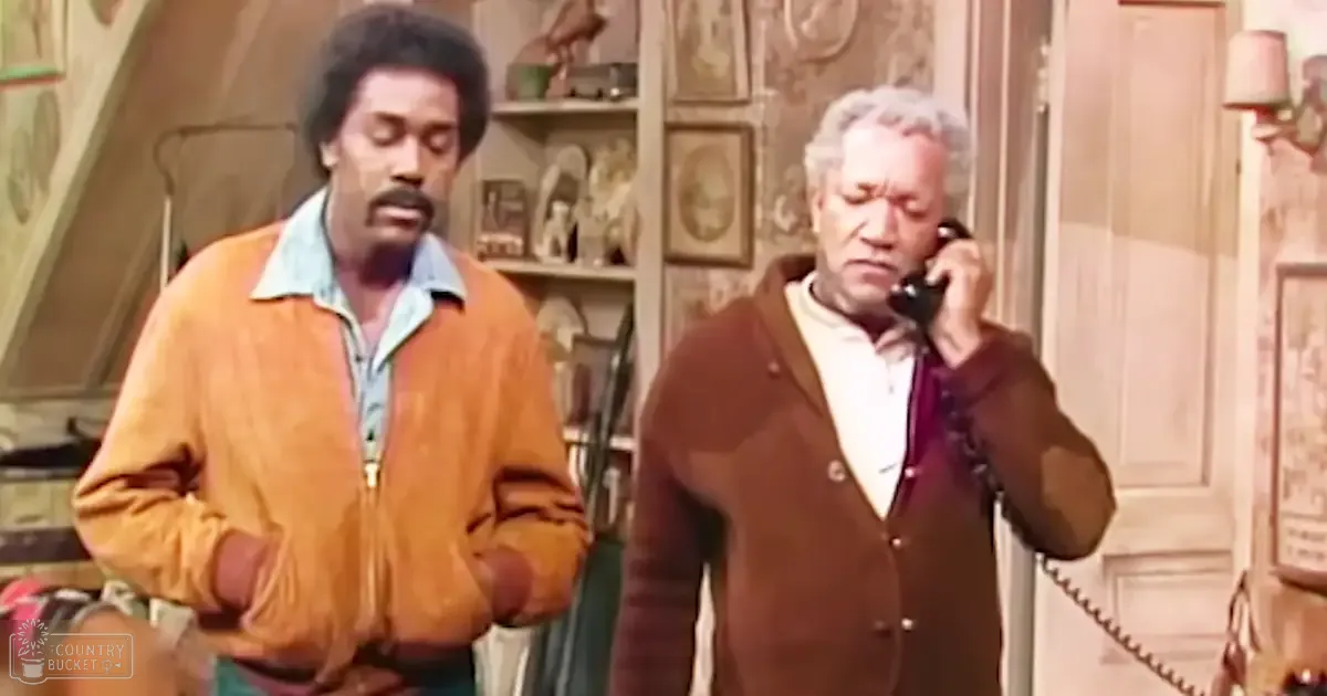 A cozy indoor home environment with two adult males, one wearing a mustard-yellow zip-up jacket and the other in a brown cardigan over white and maroon attire. The setting includes shelves of decorative items, a lamp, and framed artwork.