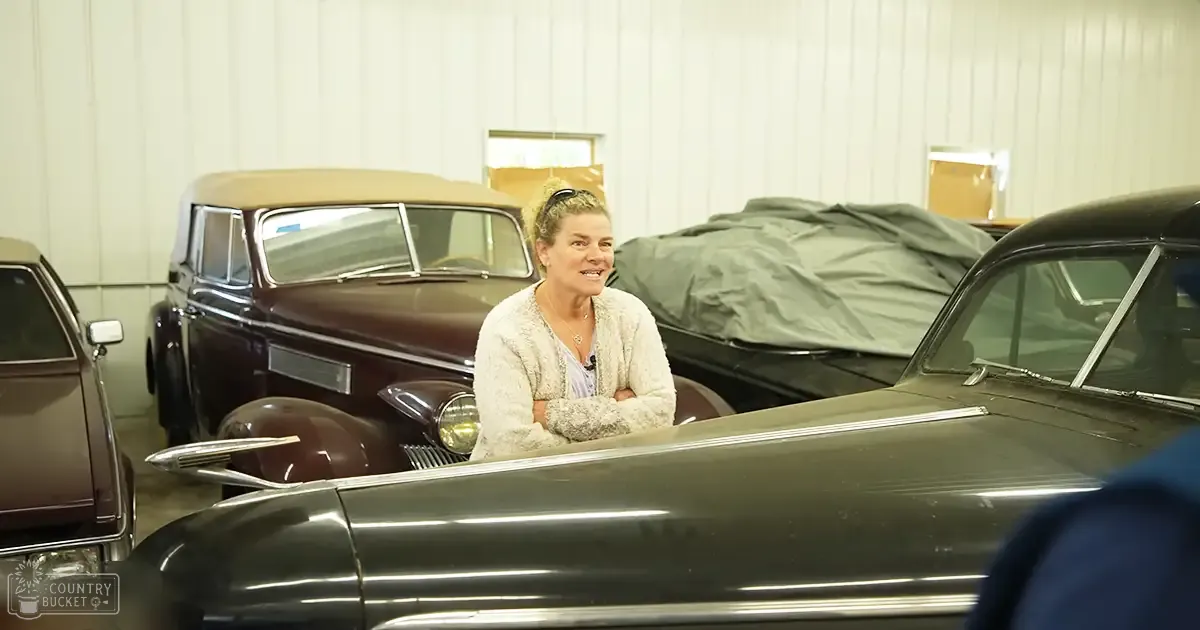 A middle-aged adult female wearing a light-colored cardigan over a patterned blouse in a vintage car showroom setting.