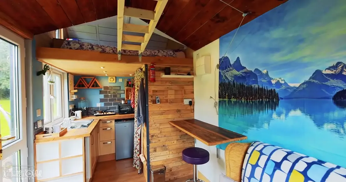 A cozy, compact living space with a wooden kitchen counter, small refrigerator, bunk bed, mural of a mountain lake scene, and natural light suggesting spring or summer.