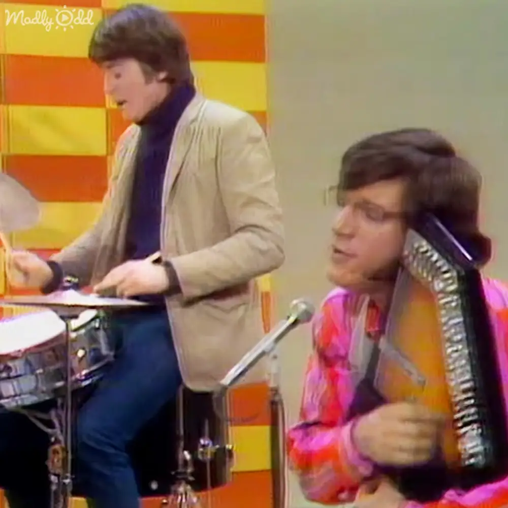 A mid-20th-century stage scene with two young adult male performers in casual attire against a brightly colored striped backdrop.