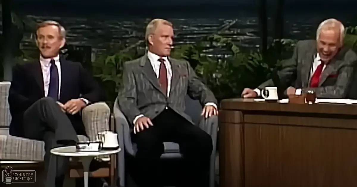Three adults seated on a talk show set with chairs, a small table between two individuals holding a mug and possibly some cards, and a larger desk where another individual is seated with a coffee cup. The background includes greenery and cityscape elements suggesting an evening setting.