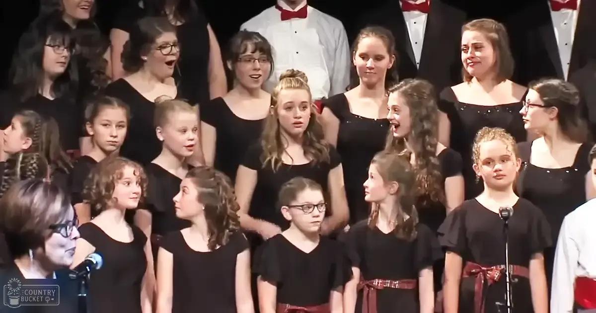 Children in black tops with red ribbons perform at a choir event under stage lighting, likely indoors during an evening performance.