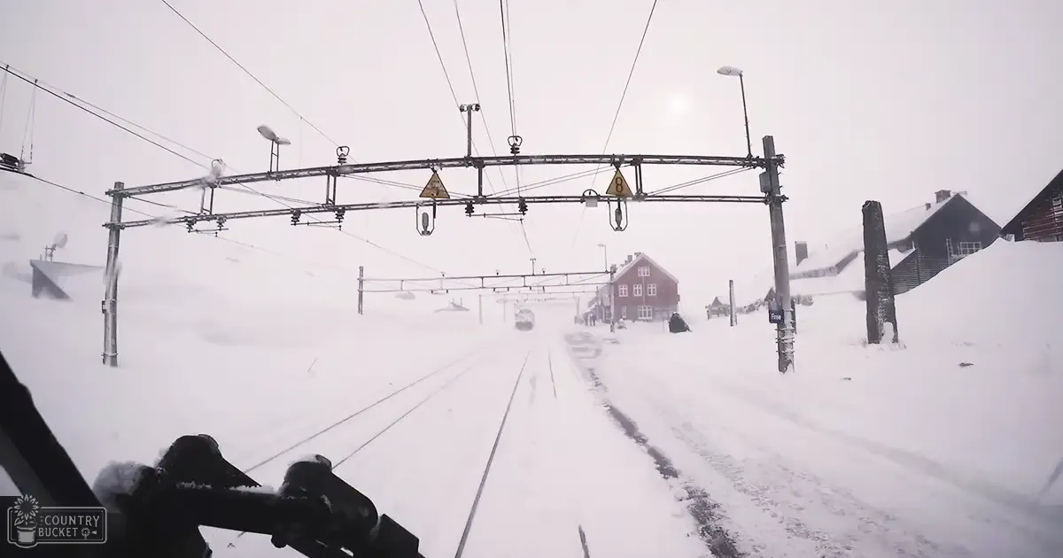 Snow-covered train station with snow-laden ground, overhead wires, signs indicating directions, and a red building in the background. Diffused light suggests early morning or late afternoon on a cold winter day.