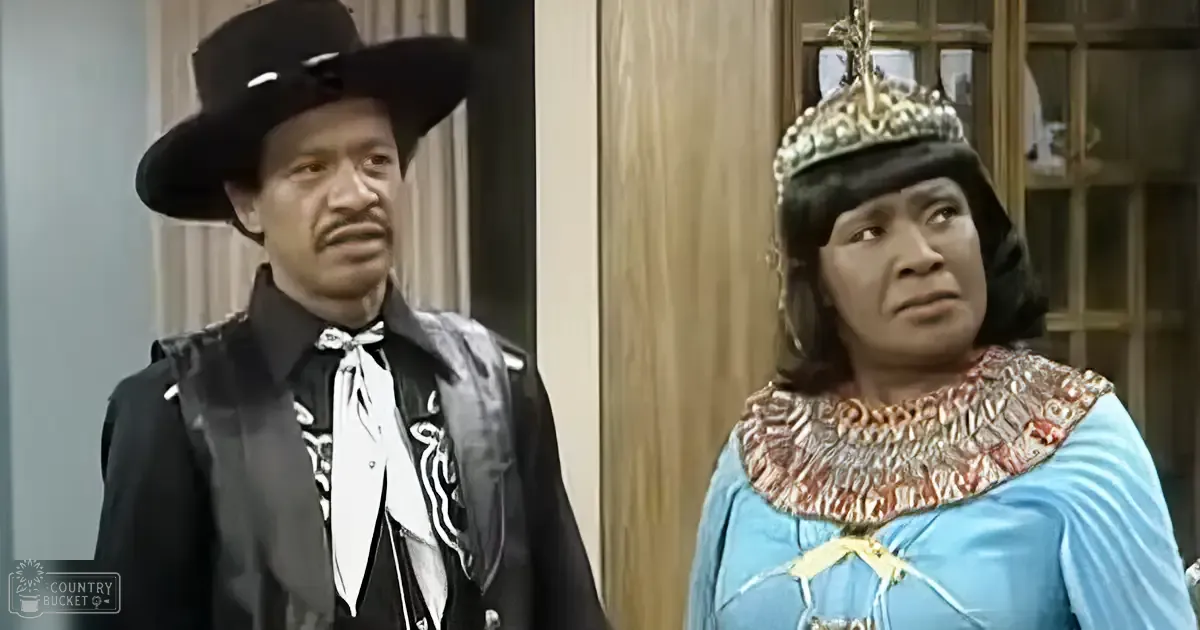 A film or TV show scene with an adult male in a black cowboy hat and vest, and an adult female wearing a blue dress with gold accents. The setting appears to be indoors with wooden paneling and glass doors.
