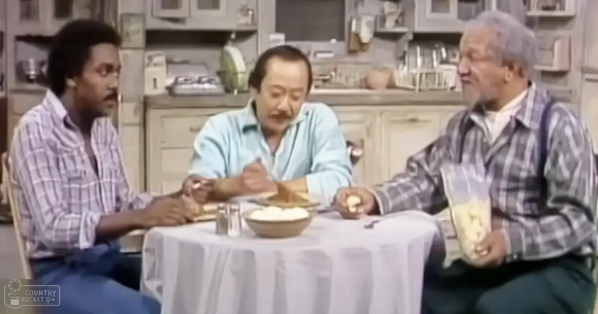 Three men wearing plaid shirts in a kitchen setting with light-colored cabinets and various kitchenware on shelves, suggesting a casual dining or cooking environment.