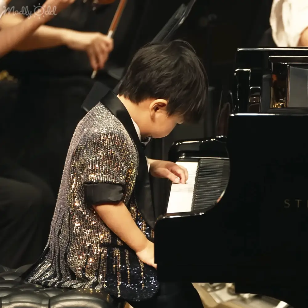 A toddler girl wearing a sparkly black dress with ruffled hem plays the piano in a performance venue, illuminated by warm evening light.