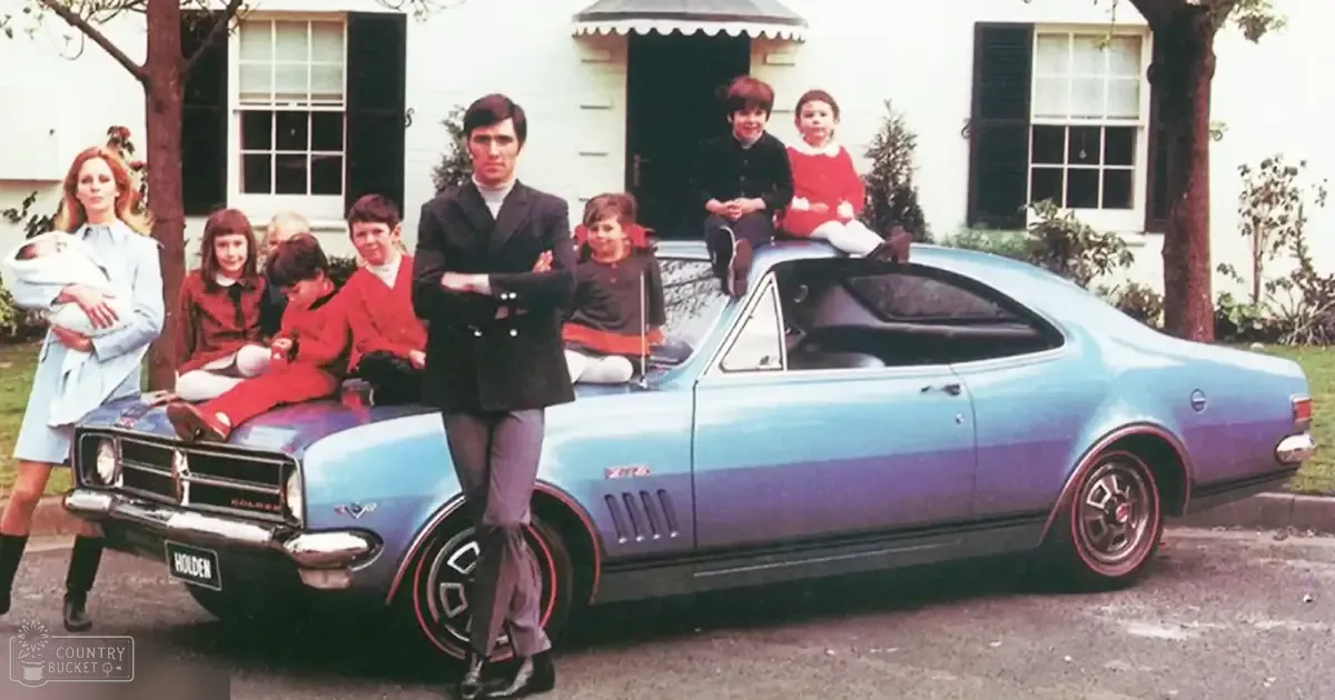 A family of adults and children sitting or standing in front of their suburban home with a vintage car parked nearby. The setting appears to be outdoors, likely spring or summer.