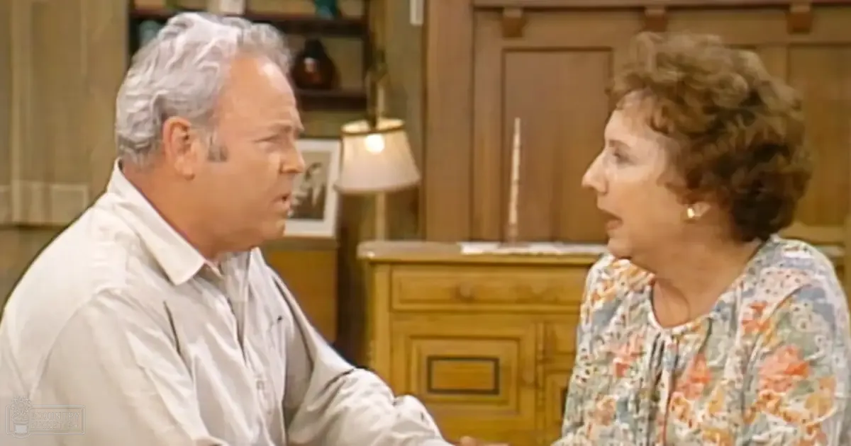 An older adult couple wearing mid-20th-century attire in a warm-lit mid-century American home interior with wooden paneling and cabinetry.