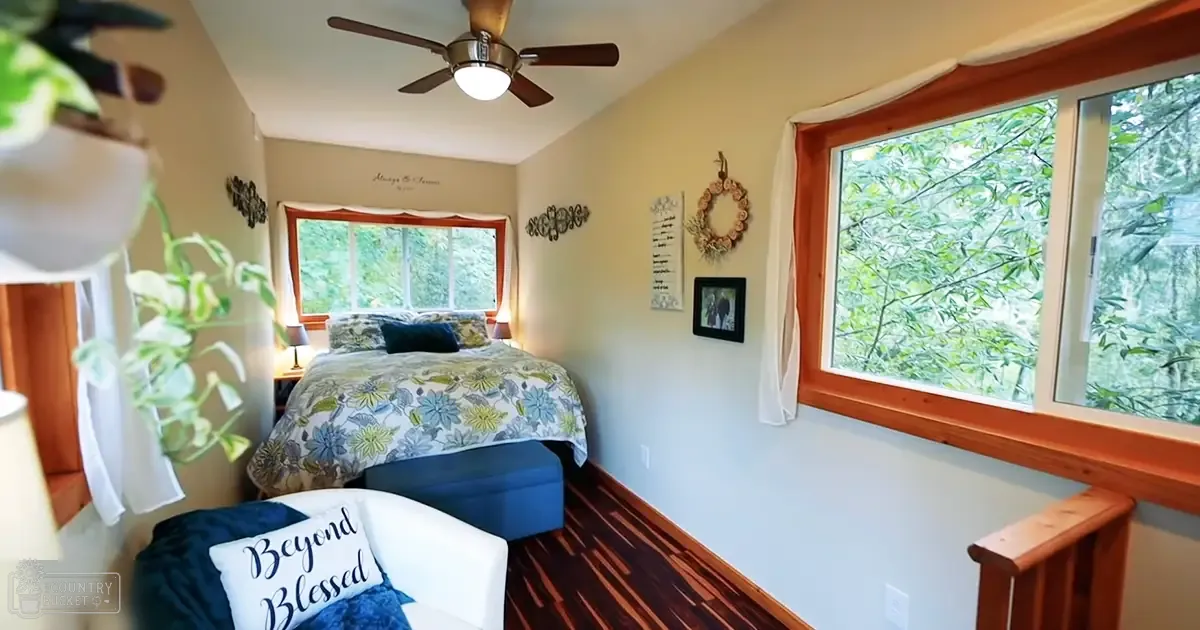 A cozy bedroom scene with floral bedding, a blue ottoman, and greenery visible through large windows in what appears to be spring or summer.