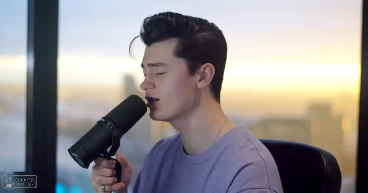 A young adult male singer wearing a light purple t-shirt is performing indoors against an urban skyline backdrop during sunrise or sunset.
