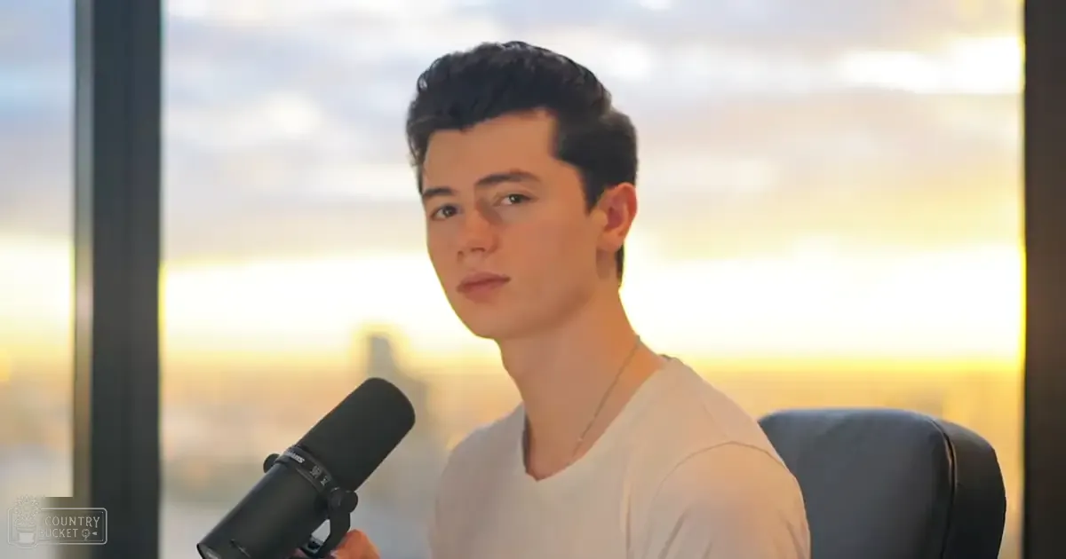 Young adult male in a relaxed fit white t-shirt with a thin chain necklace sits indoors against a scenic sunset backdrop.