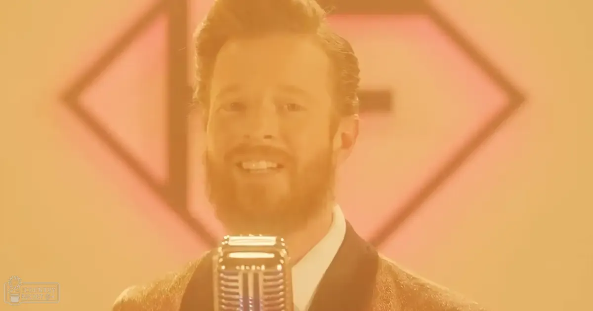 An adult male singer in a dark tailored suit with a white shirt, singing into a vintage-style microphone against an abstract geometric backdrop under warm lighting.