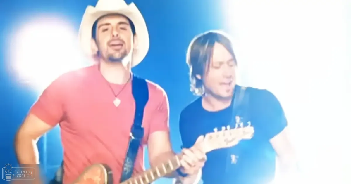 Two young adult males in casual attire perform on stage under bright blue lighting with a white cowboy hat and guitar strap visible.