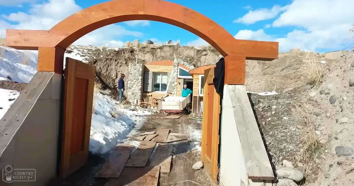 A small, partially constructed cabin in a rural setting under construction. The scene is outdoors with rocky terrain and patches of snow on the ground; the sky is clear with some clouds, suggesting late autumn or early spring.