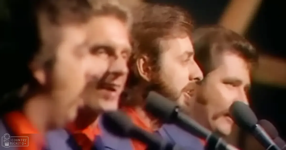 A group of adult males singing in front of microphones at an indoor event or performance, wearing dark garments with orange and red accents.