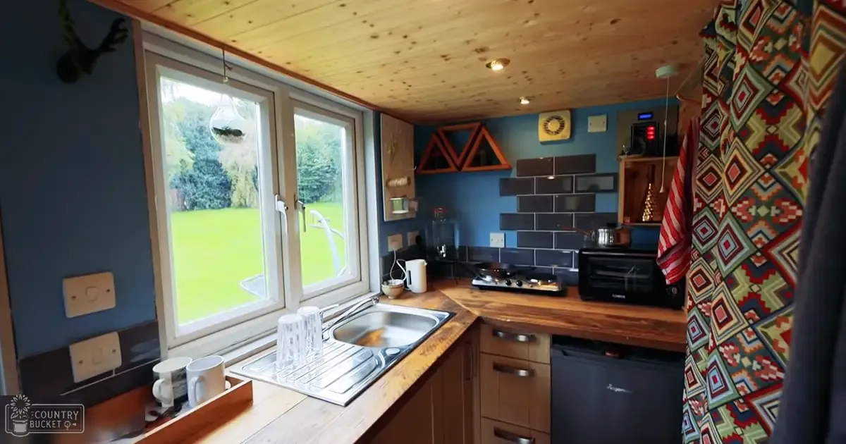 Cozy kitchen scene with stainless steel sink, wooden countertops, gas stove, microwave, colorful curtain, hanging terrarium, triangular shelf, blue walls, wooden ceiling, white-framed windows showing lush greenery, natural daylight.