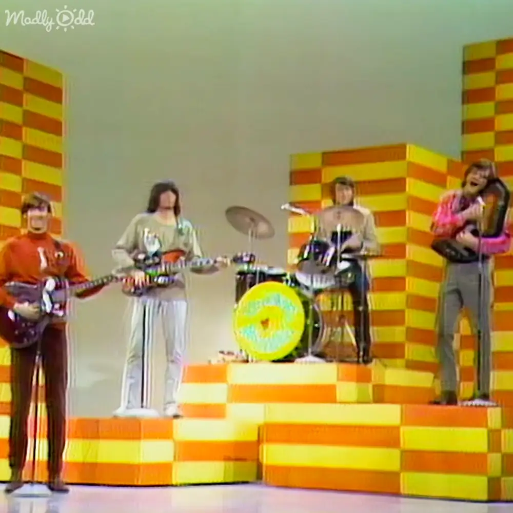 A musical performance scene with young adult males playing electric guitars and drums, an adult female holding a microphone and accordion on stage against yellow and orange striped steps under artificial lighting.
