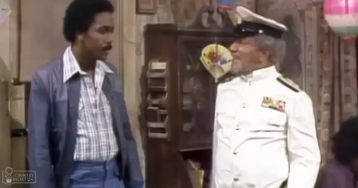 Middle-aged man wearing a blue plaid shirt with vertical stripes of white and gray, standing next to an older adult male in a white military-style uniform with epaulets and medals. They are indoors, surrounded by wooden furniture and wall decor.