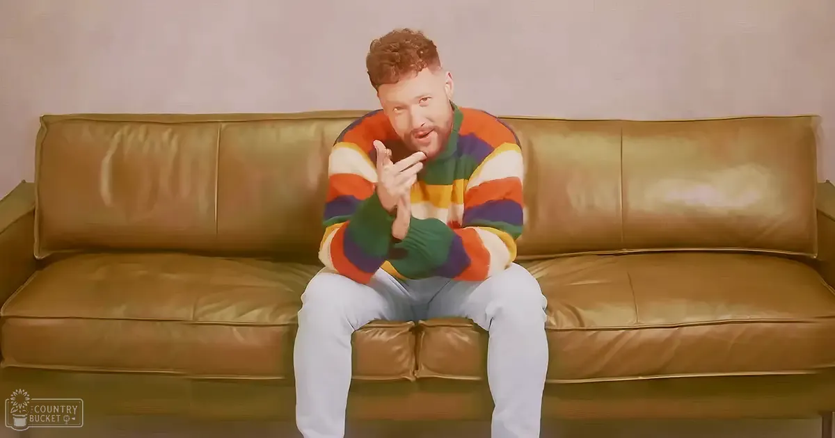 A young adult male in a colorful striped sweater sits on a brown leather couch indoors with minimal decor and soft lighting.