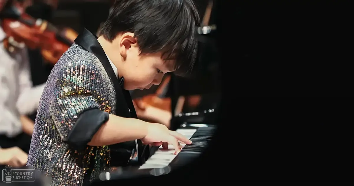 A young male child plays piano at an indoor concert, wearing a sparkly sequined jacket with black lapels and cuffs. Other musicians play violins in the background.