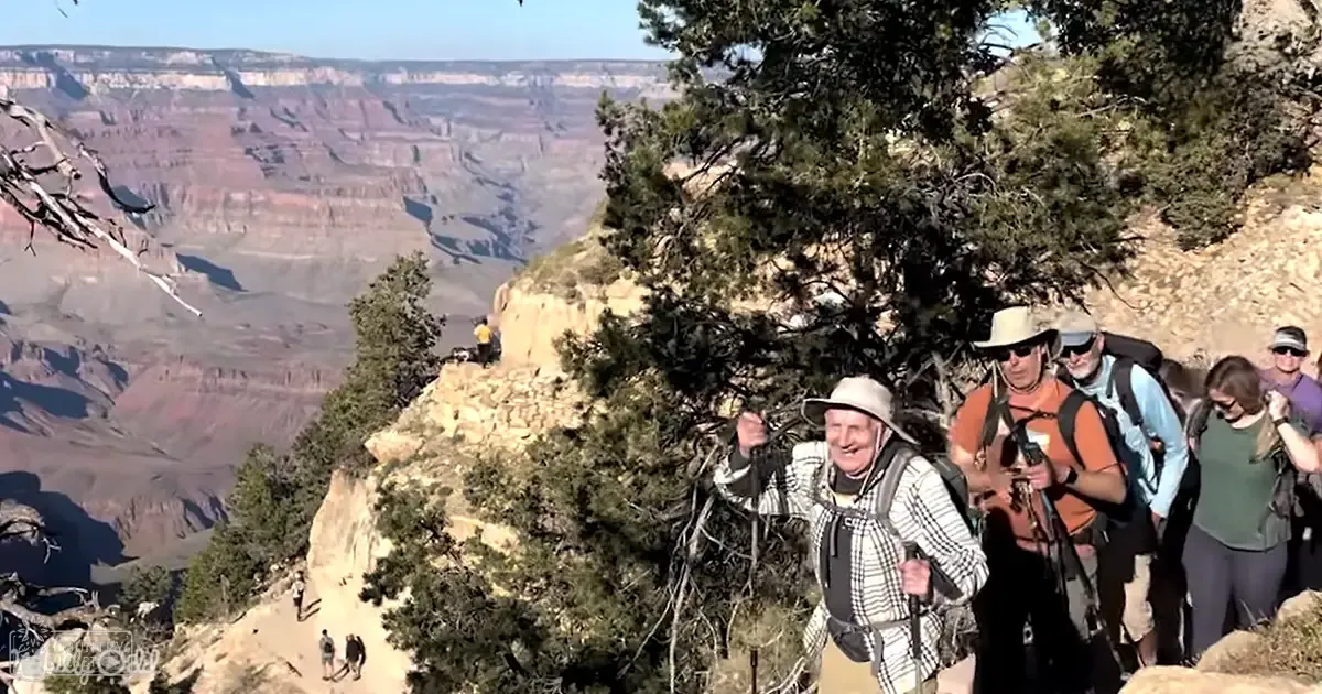 A group of hikers at the edge of the Grand Canyon, dressed in outdoor attire suitable for a spring or summer day with clear skies.