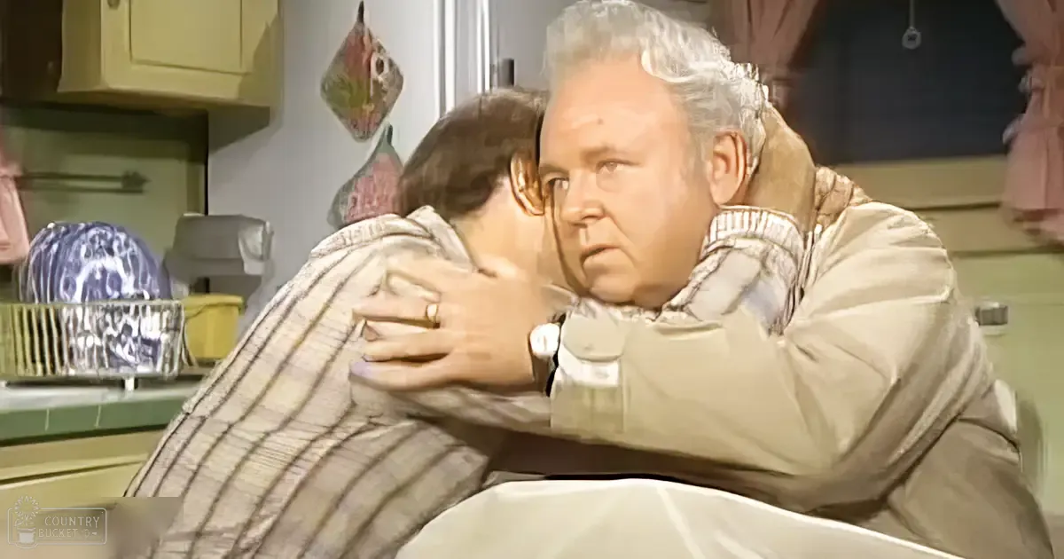 Older adult male wearing a light-colored jacket over a striped shirt, younger male in a kitchen setting with artificial lighting and various kitchen items visible.
