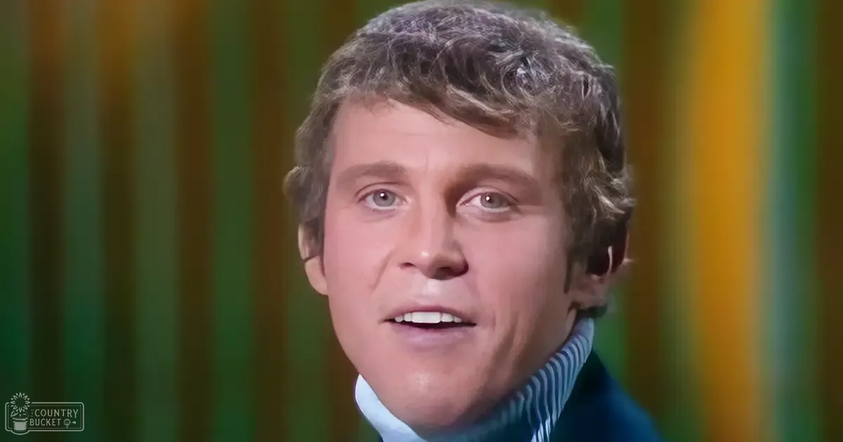 A young adult male stands indoors, wearing a blue turtleneck sweater with white horizontal stripes and a dark jacket from the mid-20th century against a backdrop resembling stage curtains in vibrant green, yellow, and orange hues.