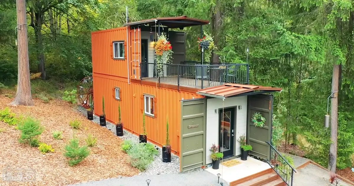 Two repurposed shipping containers stacked to form a house in a forested area with a balcony and potted plants, set outdoors during spring or summer with soft late afternoon or early evening light.
