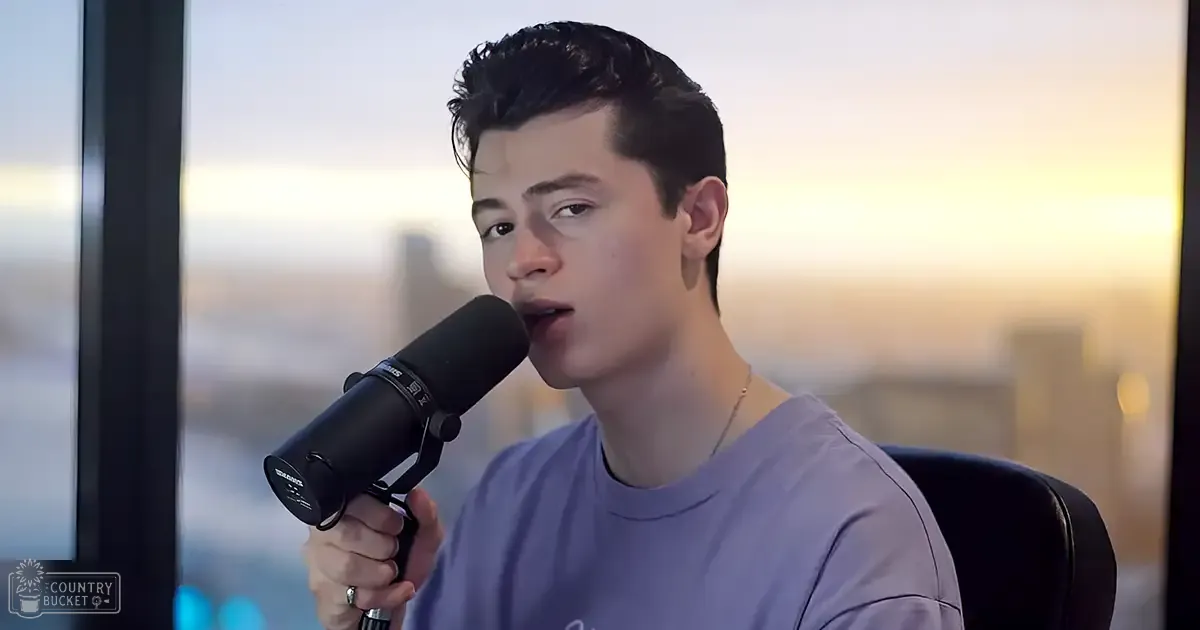 A young adult male in a relaxed fit, light purple t-shirt, holding a microphone with a pop filter indoors against a cityscape backdrop during sunset or sunrise.