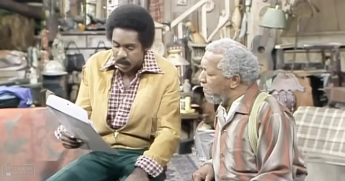 An adult male seated reading with another older adult man standing beside him in an indoor antique shop setting filled with various antiques and decorative pieces.