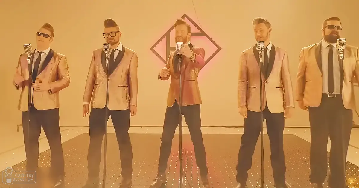A group performance scene indoors, with adult males in shiny gold blazers over white shirts with black ties, singing into microphones against a modern geometric design backdrop.
