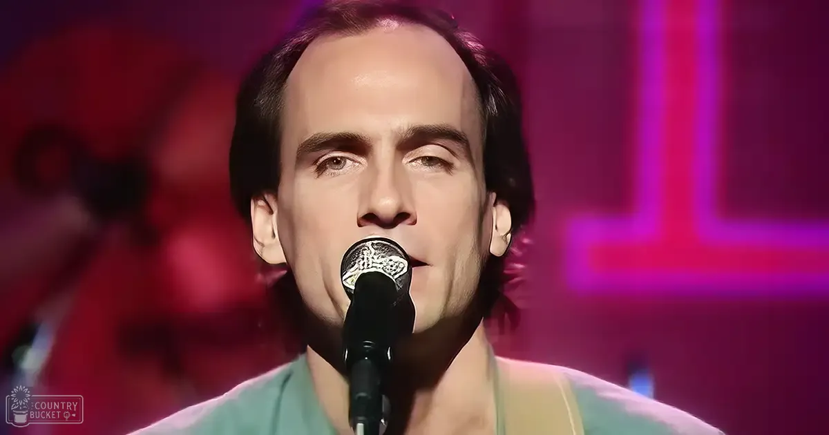 An adult male singer wearing a light green v-neck shirt, holding a microphone against his mouth in an indoor setting with red and purple stage lighting.