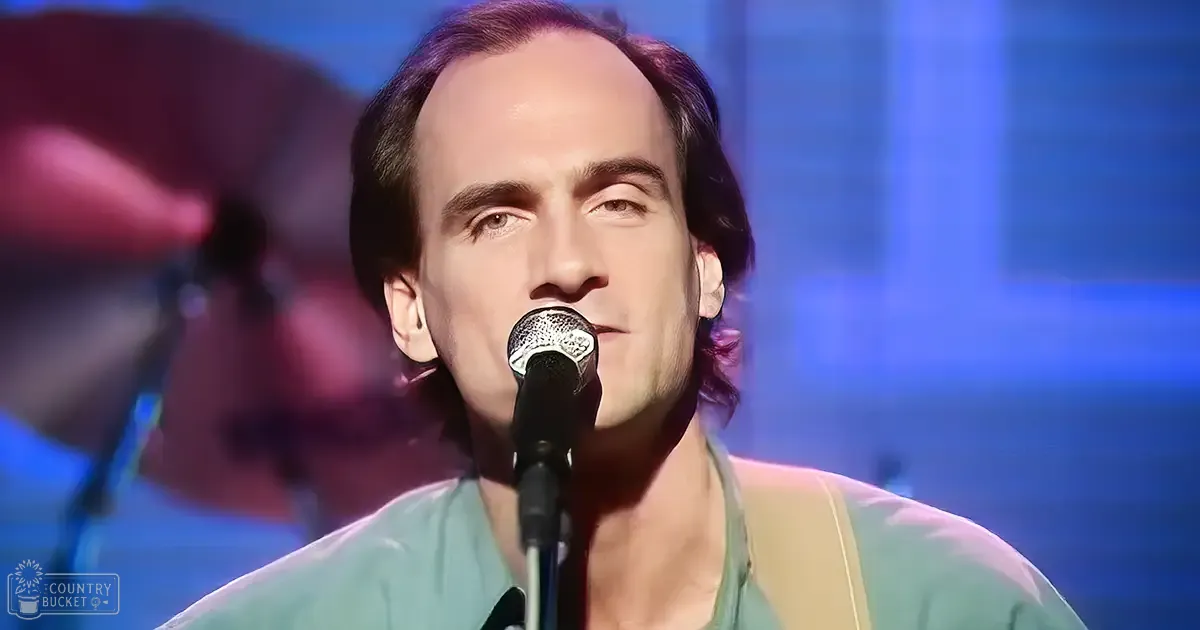 An adult male singer wearing a light green, relaxed-fit V-neck shirt with short sleeves and earplugs singing into a microphone at an indoor performance venue.