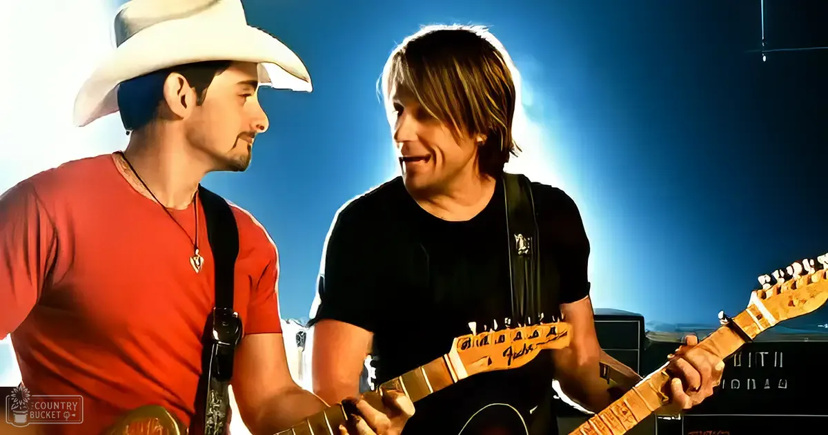Two adult male musicians play guitars on stage in a contemporary concert setting with red and black t-shirts, amplifiers, and blue stage lighting.