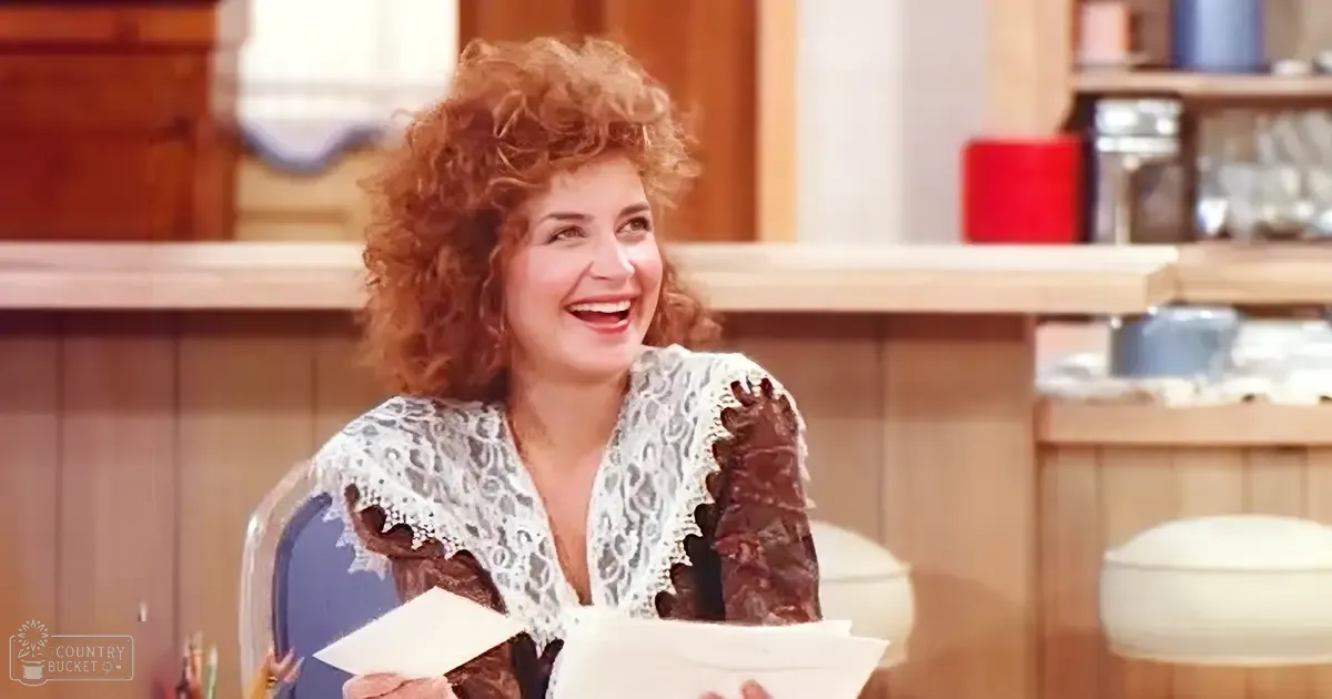 A middle-aged adult female wearing a white lace top with brown patterns and long sleeves, standing in an indoor kitchen or café setting with bright, natural lighting.