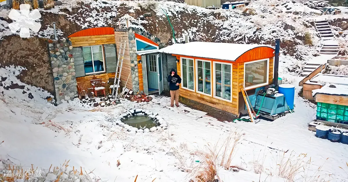 A snowy residential area with a small house built into a hillside. A person in an adult female, wearing modern casual attire and brown boots, is seen near the house.