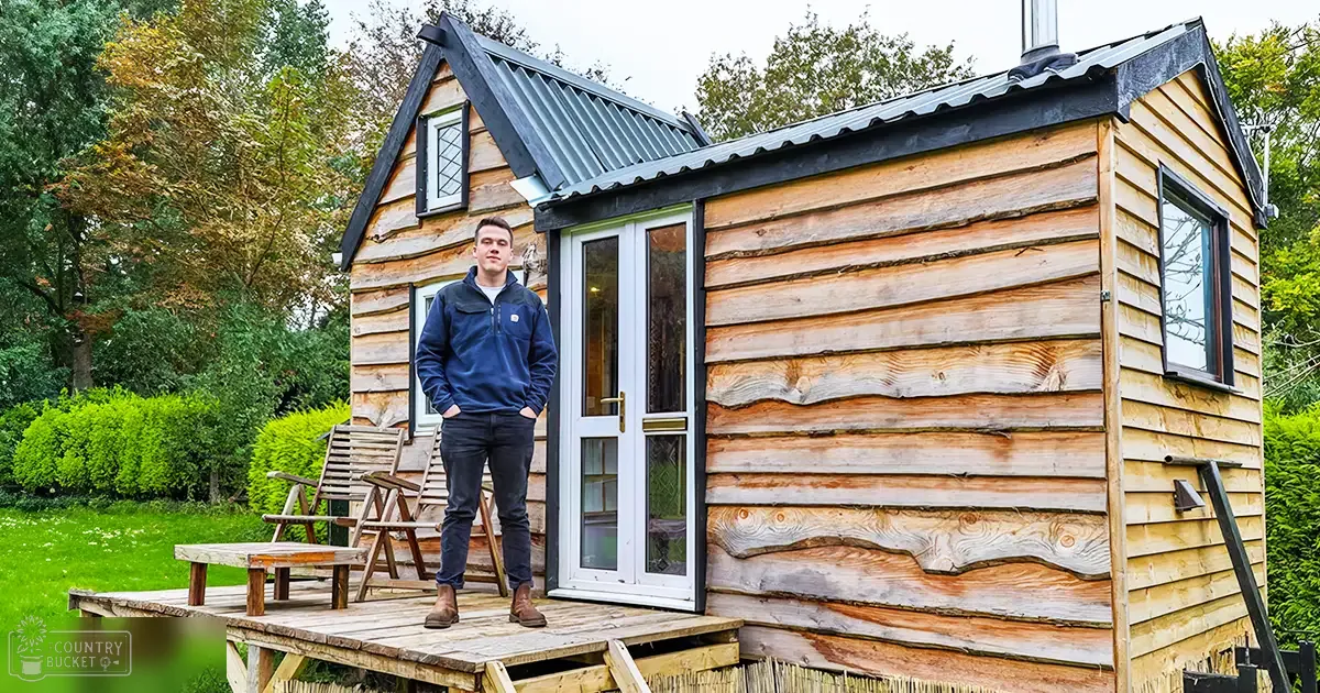 A young adult male stands in front of a small wooden cabin on an outdoor deck surrounded by greenery, suggesting late summer/early fall with soft diffused light.