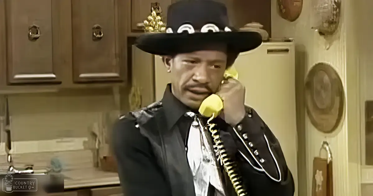 An adult male in a kitchen wearing a black leather jacket with white trim and buttons, a white shirt with patterned tie or scarf, and a wide-brimmed black hat adorned with decorative elements.