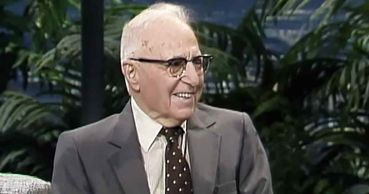 An older adult male in a studio setting, wearing a dark gray tailored suit jacket, white dress shirt, and dark brown tie with white polka dots, surrounded by lush green plants suggesting an indoor spring or summer environment.
