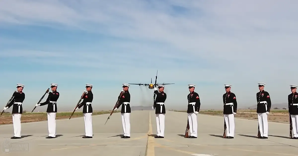 A group of young adult male military personnel in uniform standing in formation at an airfield with a parked aircraft and distant hills under clear midday light.