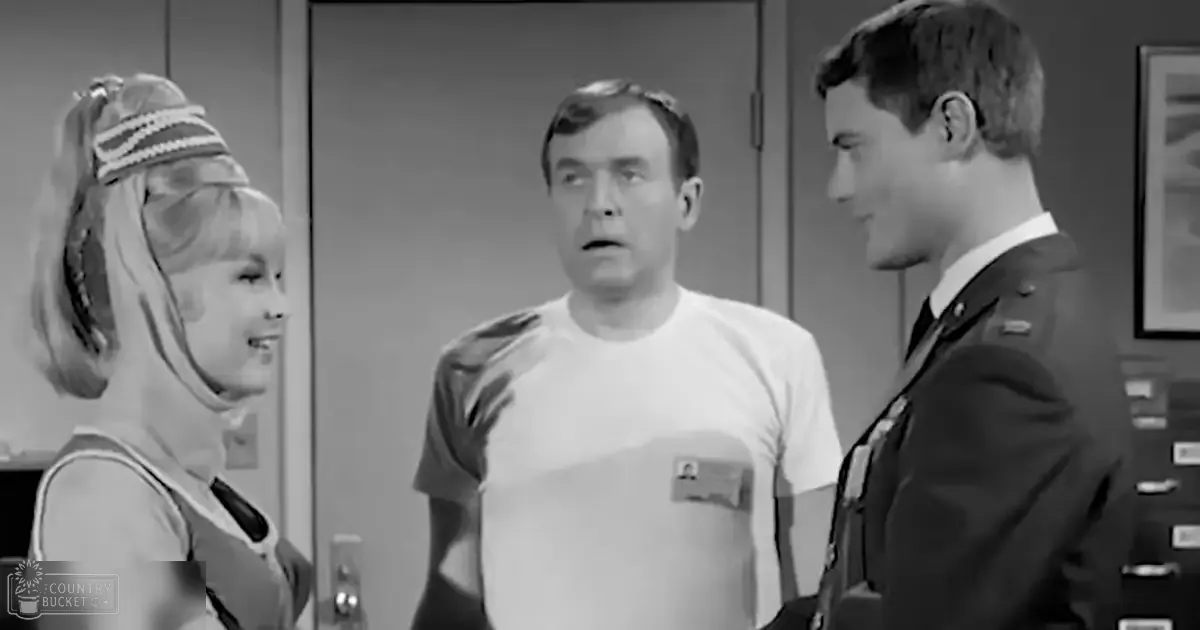 A middle-aged man in an office setting surrounded by others, including a young adult female wearing a patterned top and headband, and another male in a military-style uniform.