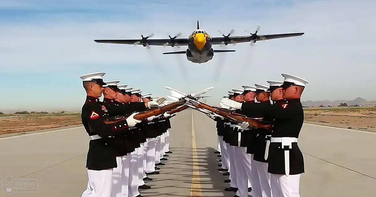 A military honor guard in dark uniforms, white trousers, and white gloves stands at attention on an airfield surrounded by a desert-like environment under natural daylight.