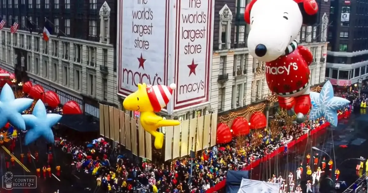 A festive parade in New York City with colorful costumes resembling Peanuts characters and holiday-themed decorations on a city street lined with tall buildings.