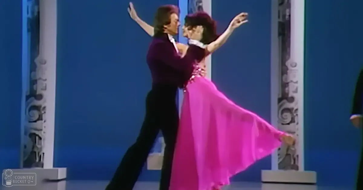 Two dancers in a studio setting, male wearing dark purple suit and white shirt, female in bright pink ballroom dress with dramatic flowing skirt, decorative columns with intricate designs in the background.